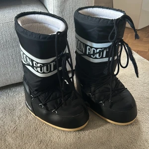 Svarta Moon Boots - Säljer ett par svarta Moon Boots i mycket bra skick. De är perfekta för vintern med sin varma och vadderade insida. Skorna har en cool design med snörning och en bred vit logga runt skaftet. Sulan är ljusbrun och ger bra grepp på snöiga underlag. Perfekta för kalla dagar! Sista bilden visar att ena snöret är lite slitet.