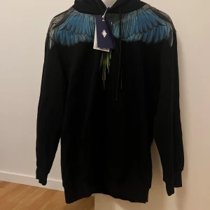 Marcelo burlon County of Milan - Säljer en svart hoodie från County of Milan med ett coolt blått vingmönster över axlarna. Den är i nytt skick med etikett kvar. Perfekt för höst och vinter, och den har en loose passform. Grym för att sticka ut i mängden!