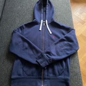 Säljer en snygg mörkblå hoodie från Ralph Lauren i storlek S. Den har en dragkedja framtill och en liten röd logga på bröstet. Tillverkad i mjuk bomull och polyester, perfekt för höst och vår. Huvan har vita snören för justering. Perfekt för en casual look! 