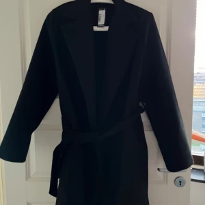 Svart kappa med bälte - Säljer en stilren svart kappa med bälte i midjan. Perfekt för höst och vår, med en elegant och tidlös design. Kappan har långa ärmar och en klassisk passform som passar de flesta tillfällen. Ett måste i garderoben för den som vill ha en chic look! Den har prislapp på och är helt oanvänd, storlek passar ej mig. 