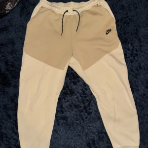 Nike Tech Fleece byxor Beige XL - Säljer ett par snygga mjukisbyxor från Nike tech byxor i beige och vitt. De har en bekväm passform med snörning i midjan och en liten Nike-logga på ena benet. Perfekta för en avslappnad stil eller träning. Byxorna har också praktiska fickor. Nypris över 1200kr! 