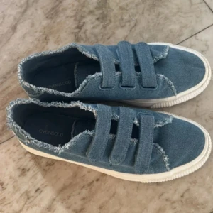 Populära jeansskor  - Säljer ett par coola blå sneakers från EVEN&ODD i jeansmaterial. De har tre praktiska kardborreband och en vit sula. Perfekta för en avslappnad stil och passar till både vår och sommar. Skorna har en fransig kant som ger en extra touch av stil.