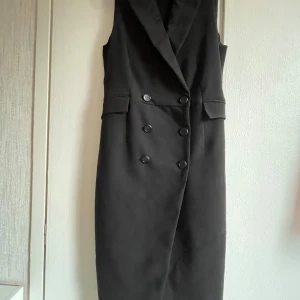 Svart ärmlös klänning med knappar - Snygg svart ärmlös klänning i normal passform med dubbelknäppning framtill. Perfekt för både fest och formella tillfällen. Klänningen har en stilren design med en klassisk krage och är knälång. Ett måste i garderoben för en elegant look!