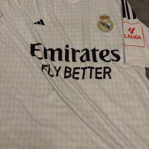 Säljer en vit Real Madrid fotbollströja i mycket bra skick. Tröjan har korta ärmar och är prydd med klubbens emblem och sponsorlogga. Den har även ett stort nummer 7 på ryggen. Perfekt för alla fans av laget!