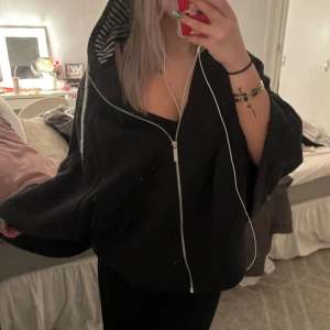 Svart hoodie med randig huva - Säljer en svart poncho med dragkedja och en cool randig insida i huvan. Den är oversized och perfekt för en chill dag eller kväll. Passar bra till både jeans och leggings. Grymt skön och stilren! Från adidas