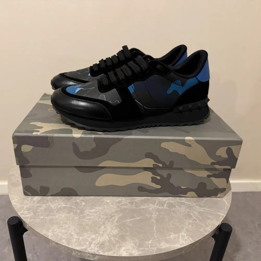 Säljer ett par snygga Valentino Rockrunner sneakers i svart och blått. Skorna har ett coolt camouflage-mönster och är i mycket bra skick. Perfekta för både vardag och fest! De har en bekväm passform med snörning och är gjorda i skinn och syntetmaterial.. Skor.