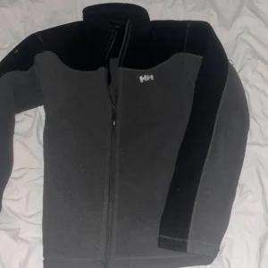 Grå fleecejacka från Helly Hansen - Säljer en grå fleecejacka från Helly Hansen i mycket bra skick. Den har en dragkedja framtill och är långärmad. Perfekt för höst och vår, och den är supermjuk och bekväm. Passar bra för både vardag och utomhusaktiviteter.