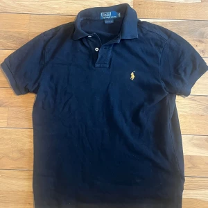 Svart pikétröja från Ralph Lauren - Säljer en klassisk svart pikétröja från Ralph Lauren med den ikoniska loggan broderad i gult på bröstet. Tröjan har korta ärmar och en bekväm passform. Perfekt för både vardag och fest!