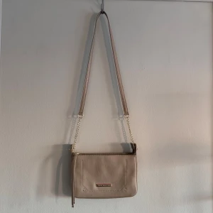 Beige axelväska från Tommy Hilfiger - Säljer en snygg beige axelväska från Tommy Hilfiger i mycket bra skick. Väskan har en stilren design med en kedjedetalj på axelremmen och en praktisk dragkedja upptill. Perfekt för både vardag och fest!