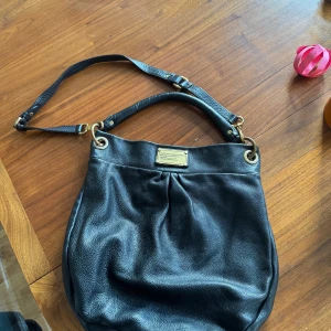 Svart axelväska Hobo bag från Marc by Marc Jacobs - Säljer en snygg svart axelväska från Marc by Marc Jacobs i skinn. Väskan har en  axelrem och en stilren design med en metallplatta framtill. Insidan är fodrad med ett mönster av bokstäver och har en praktisk dragkedjeficka. Perfekt för både vardag och fest! Insidan och metalldetaljer har spår av användningen. I övrigt fint skick. 