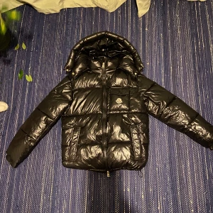 Svart dunjacka från Moncler (Montebiliard) - Säljer en snygg och varm svart dunjacka från Moncler. Jackan har en glansig finish och är perfekt för kalla vinterdagar. Den har en praktisk huva och en dragkedja framtill. Moncler-loggan sitter på bröstet och insidan har ett coolt serietryck. Perfekt för dig som vill hålla stilen i vinter! 🖤
