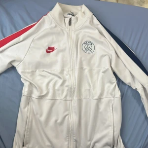 Vit träningsjacka från Nike - Säljer en snygg vit träningsjacka från Nike med PSG-logga på bröstet. Koftan har en dragkedja framtill och långa ärmar. Perfekt för sportiga aktiviteter eller som en stilren vardagsjacka. 