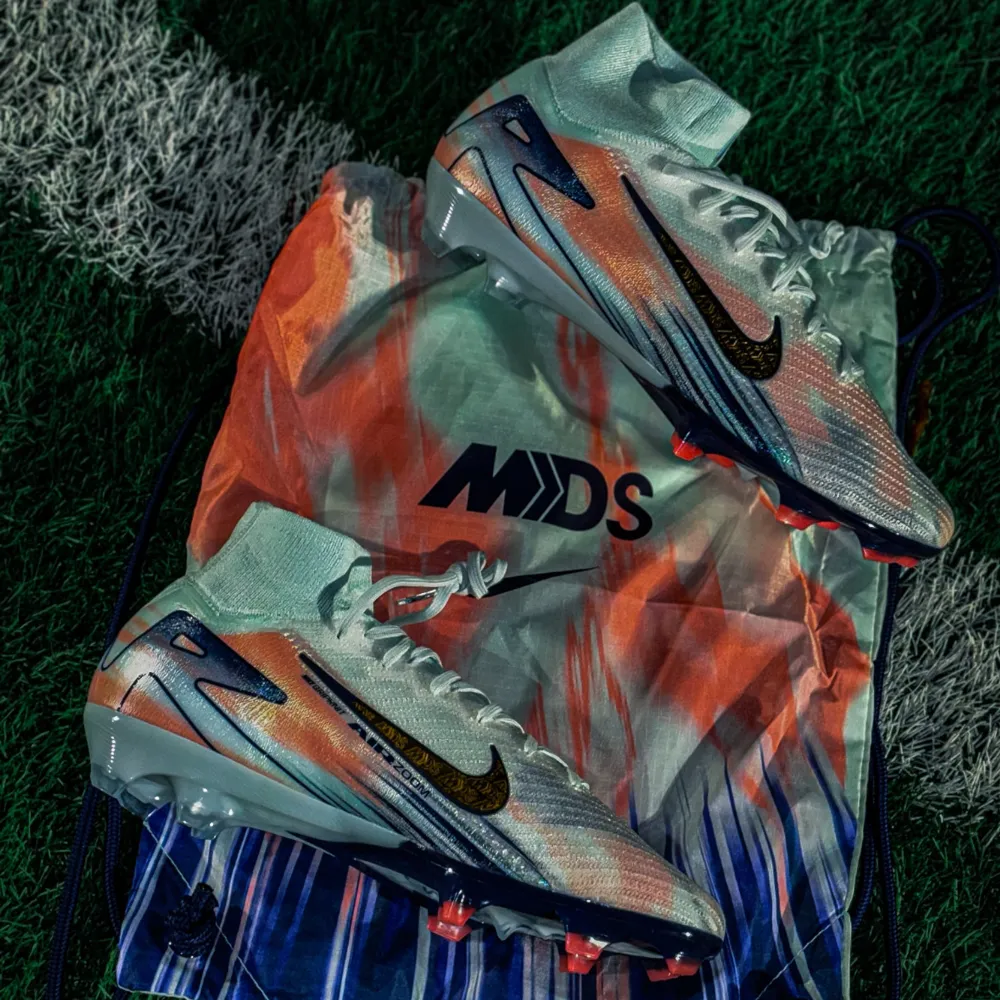 Bud tas emot!                                                         Säljer ett par Nike fotbollsskor i nytt skick. De har en cool färgkombination av vitt, blått och orange med en glittrig guld swoosh. Skorna har snörning och en hög ankel för extra stöd. Perfekta för fotbollsspelare som vill sticka ut på planen!. Skor.