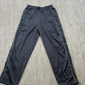 Svarta träningsbyxor med dragkedja - Säljer ett par svarta träningsbyxor i syntetmaterial. De har en bekväm loose passform med elastisk midja och snörning. Byxorna har praktiska dragkedjor vid fickorna och längs sidorna för extra ventilation. Perfekta för träning eller en avslappnad dag. Passar perfekt för hiphop, jag kör hiphop och funkar utmärkt.