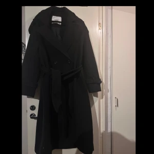 Ullkappa från Bershka - Säljer en snygg svart trenchcoat från Bershka i storlek S. Den har ett klassiskt bälte i midjan och knappar framtill. Perfekt för höst och vår, med långa ärmar och en stilren look. Passar både till vardags och festliga tillfällen!