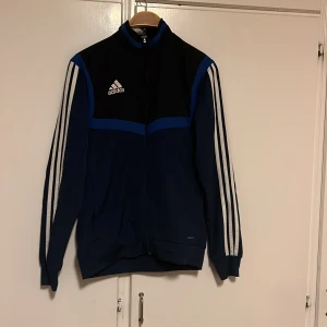 Blå och svart träningsjacka från Adidas - Säljer en snygg blå och svart träningsjacka från Adidas i storlek XS. Jackan har långa ärmar med de klassiska vita ränderna längs sidorna och en dragkedja framtill. Perfekt för träning eller vardagsbruk. Den är i bra skick och redo för nya äventyr! 🏃‍♂️