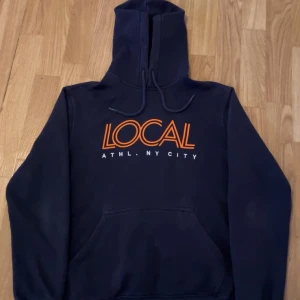 Local Hoodie - Säljer en snygg mörkblå hoodie med texten 'LOCAL ATHL. NY CITY' i orange och vitt på bröstet. Perfekt för en casual look eller att mysa i hemma. Den har en klassisk känguruficka och justerbar huva med dragsko. Passar perfekt till höst och vinter!
