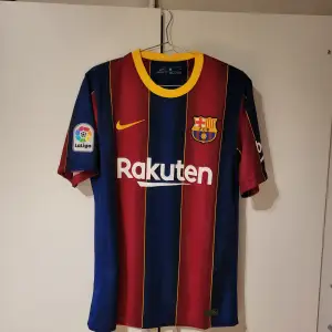 Säljer en FC Barcelona fotbollströja i mycket bra skick. Den är randig i rött och blått med gula detaljer och har korta ärmar. Tröjan är av Nike Dri-FIT material och har La Liga-märket på ärmen. Perfekt för fans av Barca! 💙❤️