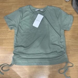 Grön ribbad topp från atmosphere - Säljer en snygg grön ribbad topp från atmosphere i nytt skick. Den har korta ärmar och en cool knytdetalj på sidorna som ger en unik look. Perfekt för vår och sommar!