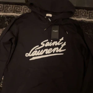 Saint Laurent hoodie - Säljer en snygg svart hoodie från Saint Laurent med deras ikoniska logga i vitt på framsidan. Perfekt för både vardag. Helt ny med taggar kvar. Passar perfekt till höst och vinter. Kvitto finns!