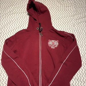 Röd hoodie med tigerbroderi från Kenzo - Säljer en snygg röd hoodie från Kenzo med ett coolt tigerbroderi på bröstet och ryggen. Den har en dragkedja framtill och långa ärmar med vita detaljer längs sidorna. Perfekt för höst och vinter, både varm och stilren. Passar lika bra till vardags som till en chill festkväll. Själva tröjan lyser även när man ute oavsett om det är ljus eller helt mörkt. Kolla bild 3&4. 