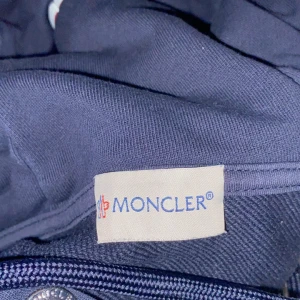 Mörkblå jacka från Moncler - Säljer en snygg mörkblå jacka från Moncler. Den har en dragkedja framtill och en huva med snörning. Moncler-loggan sitter på ärmen. Perfekt för kyliga dagar och ger en stilren look. Passar både till vardags och lite finare tillfällen.
