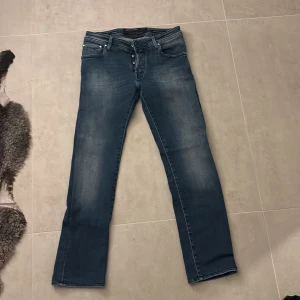 Blå jeans från Jacob Cohen - Säljer ett par snygga blå jeans från Jacob Cohen i bra skick. De har en klassisk femficksdesign och knappar framtill. Jeansen har en stilren look med en liten sliten effekt för en cool touch. Perfekta för både vardag och fest               Nypris: 5000kr