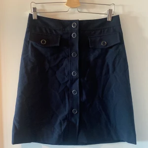 Mörkblå kjol från Filippa K - Snygg mörkblå kjol från Filippa K i mycket bra skick. Kjolen har en rad med knappar framtill och dekorativa fickor. Perfekt för både vardag och fest! 