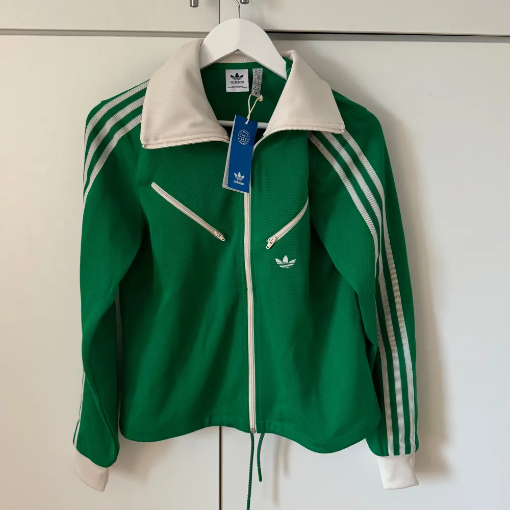 Säljer en snygg grön träningsjacka från Adidas med vita detaljer. Jackan har dragkedja framtill och två dragkedjefickor på bröstet. Den har långa ärmar med de klassiska tre ränderna längs sidorna och en vit krage. Perfekt för träning eller en sportig vardagslook!. Jackor.