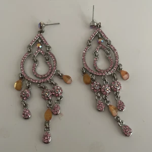 Glittriga örhängen med rosa och orange detaljer - Säljer ett par fantastiska örhängen med glittriga rosa stenar och orange droppformade detaljer.Två stenar på ena örhänge saknas. De har en elegant design med flera hängande delar som ger en fin rörelse. Perfekta för att lysa upp vilken outfit som helst! 🥰