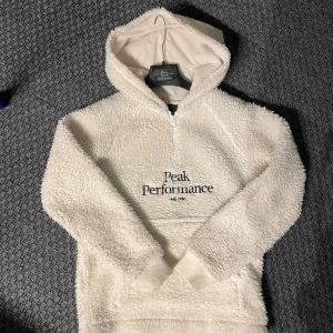 Teddy hoodie från Peak Performance - Mysig vit Teddy hoodie från Peak Performance. Den har en halv dragkedja och en stor ficka framtill. Superbekväm och varm med en fluffig textur. Perfekt för både vardag och utomhusaktiviteter.