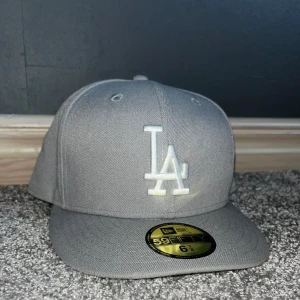 Grå keps från New Era - Säljer en stilren grå keps från New Era, modell 59FIFTY. Kepsen har en broderad vit LA-logga på framsidan och är i nytt skick. Perfekt för både vardag och sportiga aktiviteter. Passar bra till alla årstider pris kan diskuteras vid snabb affär 🤩🤩