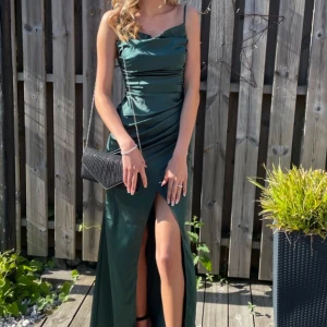 Mörkgrön balklänning med slits - Säljer en elegant mörkgrön långklänning i satinliknande material med en stilren slits framtill. Klänningen har smala axelband och en figurnära passform som framhäver silhuetten. Endast använd 1 gång så i superskick då det inte finns någon skada. 