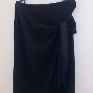 Svart kjol med knyt - Snygg svart kjol med knyt framtill som ger en elegant touch. Perfekt för både vardag och festliga tillfällen. Kjolen har en omlott-design som gör den extra chic och stilren. Passar bra till både sommar och höst. Ett måste i garderoben för den som älskar klassiska plagg!