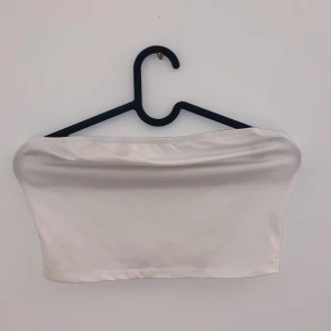 Vit axelbandslös tubtopp - Säljer en stilren vit axelbandslös tubtopp i mjukt material. Perfekt för sommardagar eller som basplagg under en kavaj. Toppen är i bra skick och passar till många olika outfits. Perfekt för både vardag och fest!