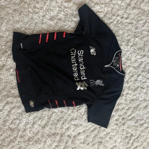 Svart fotbollströja från New Balance - Säljer en svart fotbollströja från New Balance med korta ärmar. Den har röda detaljer på sidorna och ett tryck av 'Standard Chartered' på framsidan. Perfekt för fotbollsfans som vill ha en stilren och bekväm tröja. Passar bra till både träning och vardag. ÄKTA!