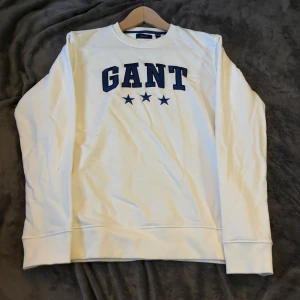 Vit sweatshirt från GANT - Säljer en stilren vit sweatshirt från GANT med blå text och stjärnor på framsidan. Tröjan har långa ärmar och är i mycket bra skick. Perfekt för en avslappnad stil eller vardagsbruk. Passar både höst och vår.