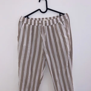 Randiga linnebyxor - Säljer ett par snygga randiga linnebyxor i beige och vit. De har en avslappnad passform med resår och snörning i midjan för extra komfort. Perfekta för sommardagar eller en avslappnad vårlook. Byxorna är i bra skick och redo för nya äventyr!