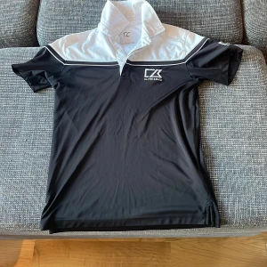 Svart och vit pikétröja från Cutter & Buck - Säljer en stilren svart och vit pikétröja från Cutter & Buck i storlek S. Tröjan har korta ärmar och en klassisk krage med knappar. Perfekt för golf eller en avslappnad dag ute. Materialet känns lätt och bekvämt. Passa på att köpa denna snygga tröja! 🖤🤍Pris går alltid att förhandlas!