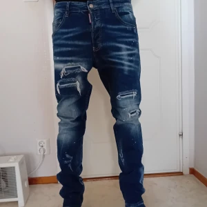 Blå jeans från Dsquared2 - Säljer ett par snygga blå jeans från Dsquared2 i bra skick. De har en cool slitningseffekt och färg stänk ,knappar framtill. Perfekta för en avslappnad stil med en touch av edge. Passar både till vardags och fest! Perfekt nu till vintern. Stolek 50 och waist 34/35. Kommer från ett rök och djur fritt hem.