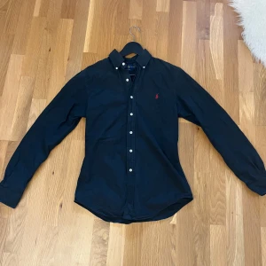 Svart skjorta från Ralph Lauren - Säljer en stilren svart skjorta från Ralph Lauren i slim fit. Skjortan har långa ärmar och knappar fram. Den är i mycket bra skick och perfekt för både vardag och fest. Den klassiska designen gör den lätt att matcha med olika outfits. Passar perfekt för höst och vår!