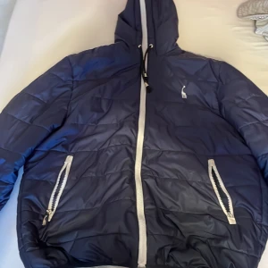 Mörkblå pufferjacka med huva - Säljer en snygg mörkblå pufferjacka med huva. Jackan har en vit dragkedja och praktiska fickor med dragkedjor. Perfekt för kyliga dagar och håller dig varm och bekväm. Passar både till vardags och lite mer avslappnade tillfällen. 🧥