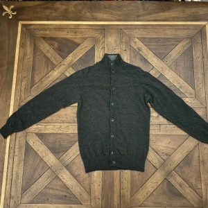 Grå kofta från Hackett - Säljer en elegant grå kofta från Hackett som passar S/M. Den är tillverkad i en lyxig blandning av merinoull, silke och kashmir, vilket gör den supermjuk och bekväm. Koftan har långa ärmar och knappar i ben framtill. Perfekt för höst och vinter!