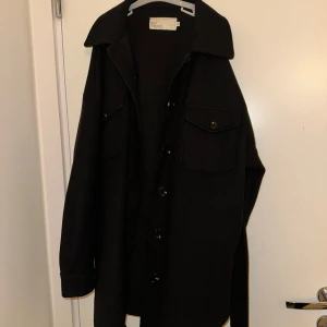 Svart kappa från Nelly - Snygg svart kappa från Nelly i mycket bra skick. Den har långa ärmar och knappar framtill samt ett bälte i midjan för en stilren look. Perfekt för höst och vår!