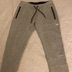 Grå mjukisbyxor från Jack & Jones - Säljer ett par grå mjukisbyxor från Jack & Jones i bra skick. De har en bekväm passform med snörning i midjan och praktiska dragkedjor på fickorna. Perfekta för en avslappnad dag hemma eller lätt träning. Materialet är en blandning av polyester och bomull vilket gör dem mjuka och hållbara.