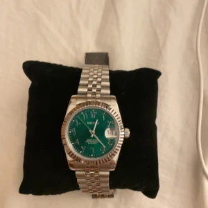 Seiko Mod Grön Arabic - Säljer en stilren klocka med en grön urtavla och datumvisning. Klockan har ett silverfärgat armband i metall och en klassisk design. Perfekt för både vardag och fest! Datumen ändras kl 03:00. Extra länkar följer med och ändras på Sjärnmurmakarna har kvitto hos dem som medföljer i köp. Samt en klocka låda 