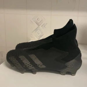 Svarta fotbollsskor från Adidas - Säljer ett par svarta fotbollsskor från Adidas i mycket bra skick. Skorna har en tight passform och är perfekta för fotbollsträning. De har en cool design med grå detaljer och är gjorda i syntetmaterial för bästa hållbarhet och prestanda på planen.