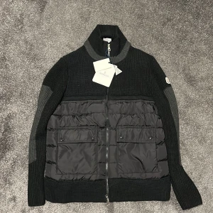 Svart stickad jacka från Moncler - Säljer en snygg svart stickad jacka från Moncler i nytt skick. Den har en dragkedja framtill och quiltade detaljer på framsidan. Perfekt för höst och vinter med sin varma och stilrena design. Storlek M.