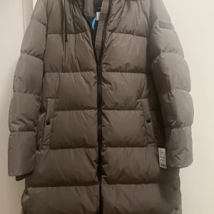 Brun dunjacka från Rockandblue - Säljer en supermysig brun dunjacka från Rockandblue i storlek 42. Perfekt för kalla vinterdagar med sin långa design och varma vaddering. Jackan har en dragkedja framtill och långa ärmar. Den är i nytt skick och gjord av återvunnen polyester. En riktig vinterfavorit! ❄️