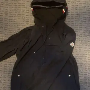 Säljer en snygg svart jacka från Moncler. Den har en praktisk huva och dragkedja framtill. Perfekt för höst och vår, med en stilren design och Moncler-logga på ärmen. Jackan har långa ärmar och knappar vid fickorna. Passar både till vardags och lite finare tillfällen. Passar M också!